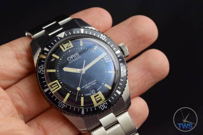 The Oris Divers Sixty-Five (With Metal Bracelet) [01 733 7707 4064-07 8 20 18] held in the hand with a black bacground